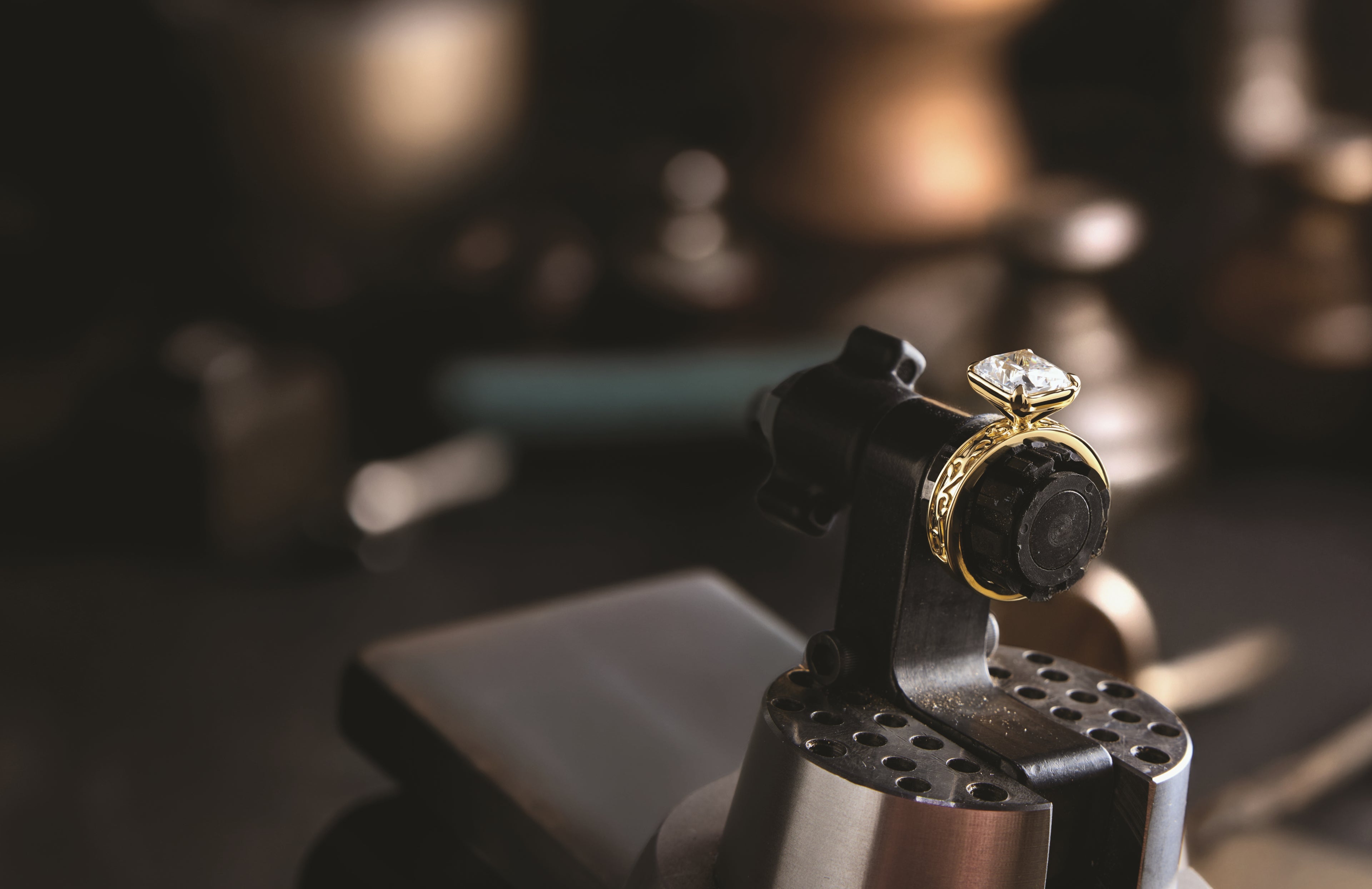 A gold ring with a clear gemstone displayed on a jeweler's stand in a dark, softly blurred workshop setting.