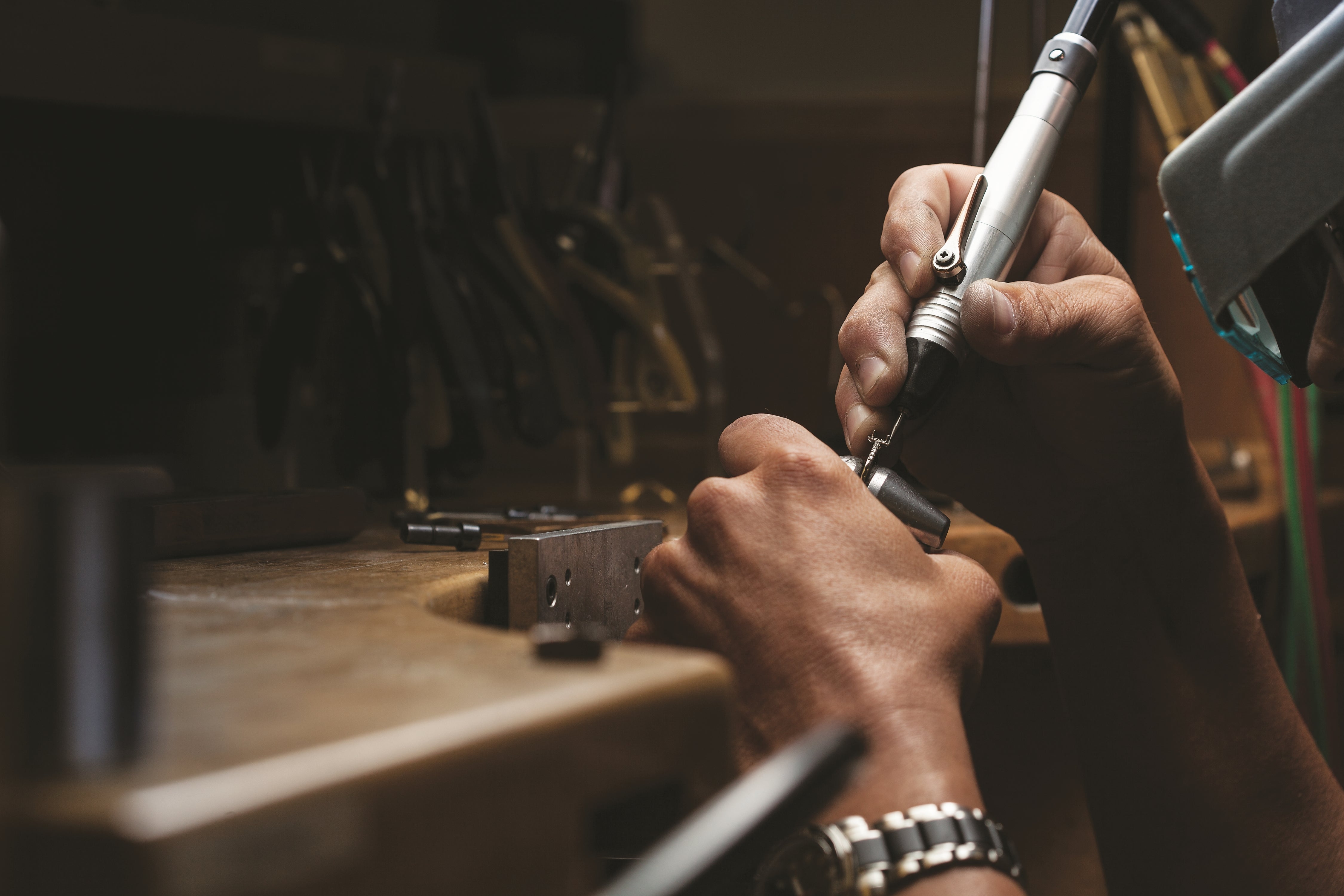 Jeweler working at a bench, using a handheld rotary tool to detail a piece of jewelry.
