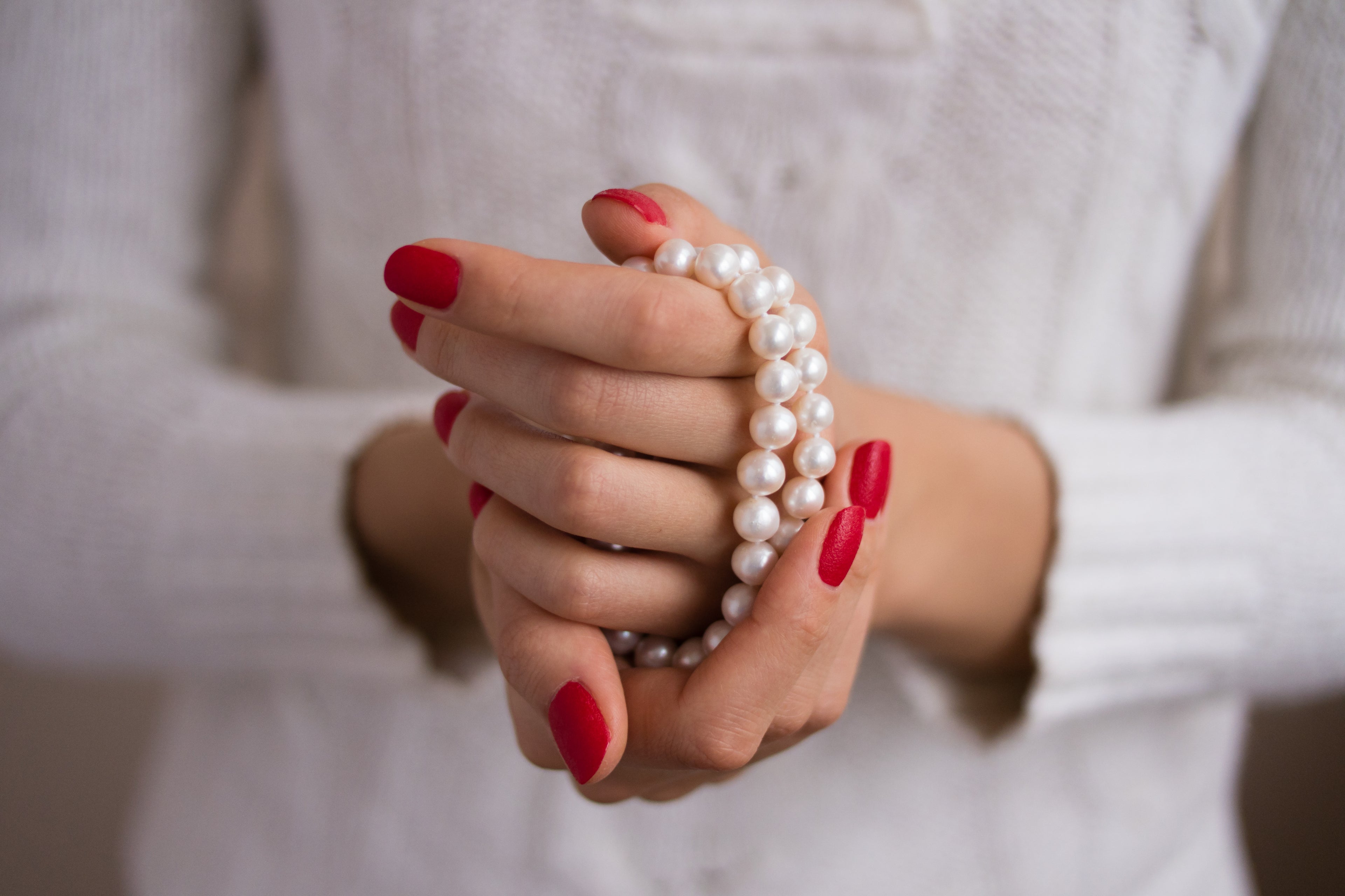 A close-up of a person's hands with red nail polish holding a white pearl strand bracelet against a white knit sweater.